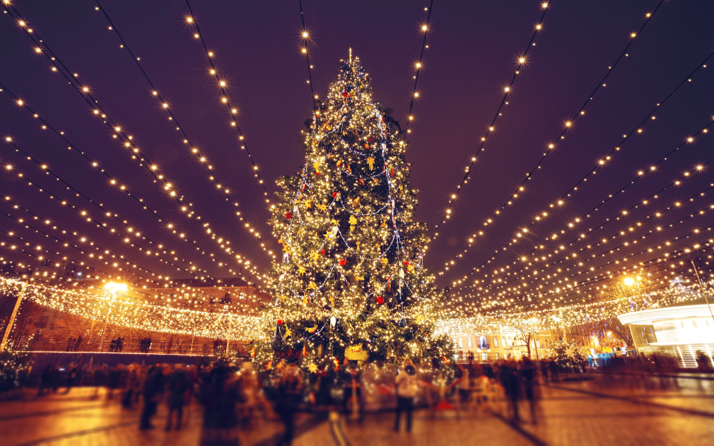 Large Christmas tree decorated with lights and ornaments in a public square, surrounded by a canopy of festive string lights at night.