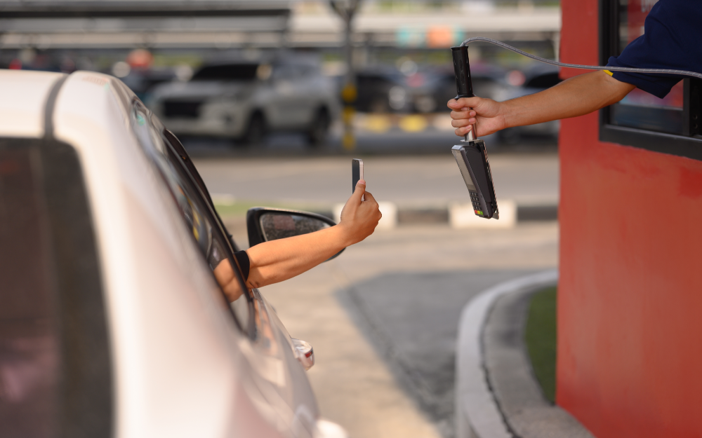 A car at a drive-thru window at a fast food, a customer is using a mobile phone while the cashier holds out a card reader.